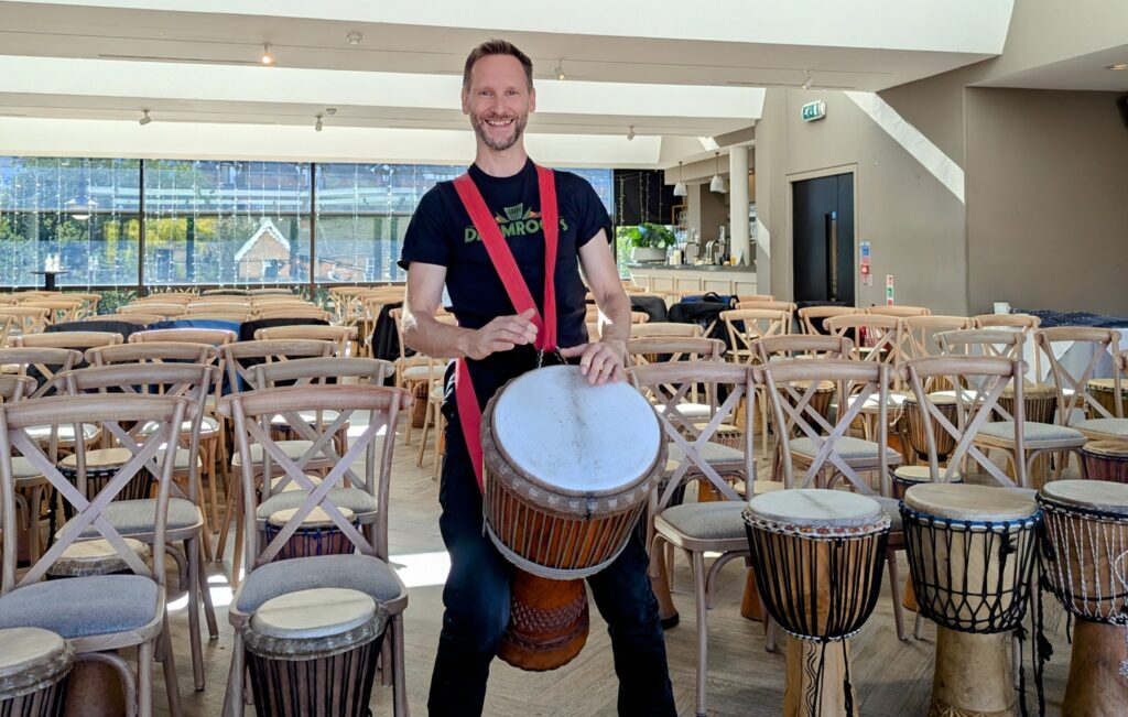 A man standing with an African drum held with a harnedd in front of many African drums in front of chairs