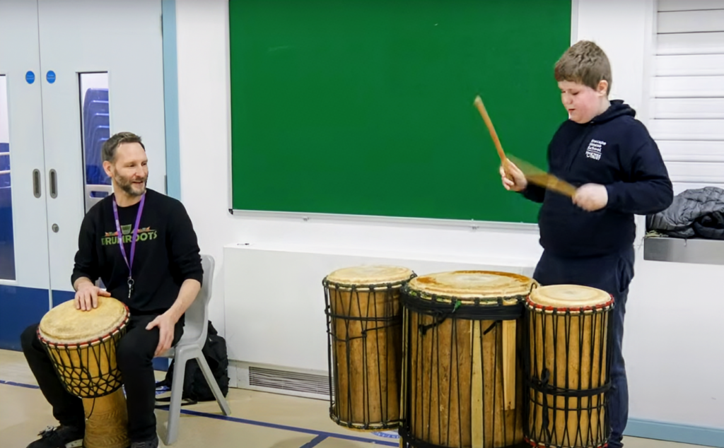 A man sitting with an African djembe drum watching a Boy playing three large African drums with drumsticks