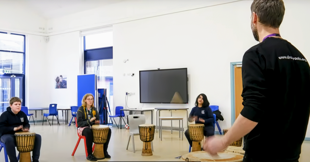 A man standing playing African drums with sticks whilst 3 students play African djembe drums sitting on chairs.