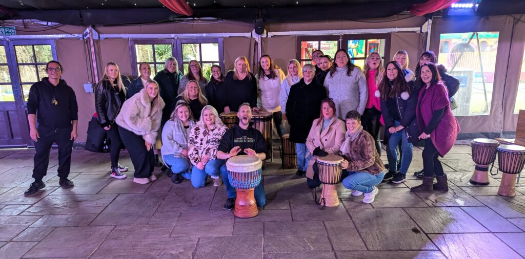 A group of about 20 people mostly women posing for a group photo with a few African drums in the foreground